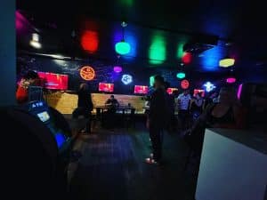 A dimly lit bar with neon lights on the walls and colorful ceiling lights, located in Uptown Waterloo. People are standing and sitting at tables, some chatting while others explore a few screens mounted on the walls. An ATM is visible in the foreground. The atmosphere appears casual and lively.