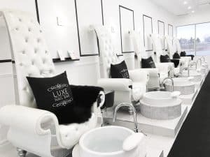A chic nail salon in Uptown Waterloo features plush white high-back chairs with armrests, each accompanied by a black throw pillow and a matching black blanket. Below each chair is a white foot basin for pedicures. The room is illuminated by natural light from large windows on the right, inviting you to explore Uptown's elegance.