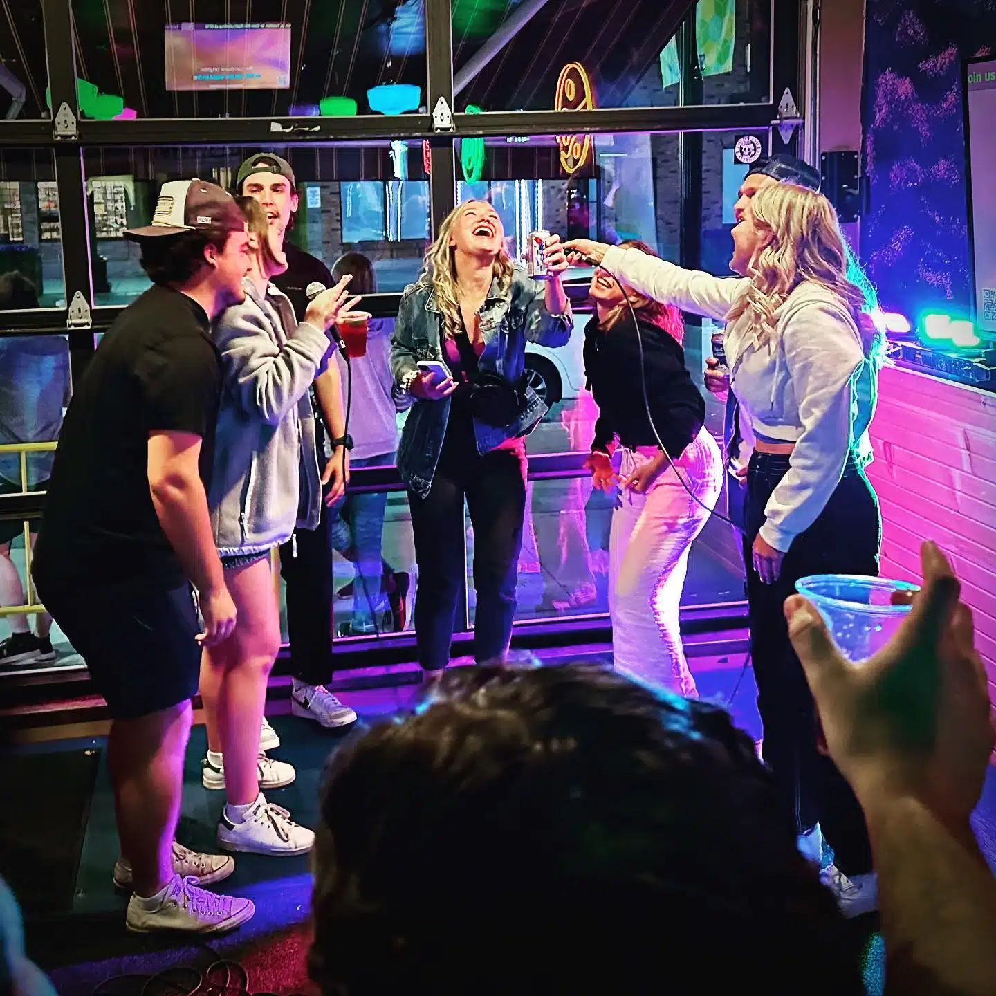 A group of six people are gathered together in a lively indoor setting in Uptown Waterloo, where one person holds a microphone and the rest are laughing and cheering. The scene is vibrantly lit with colorful lights, creating an energetic and fun atmosphere perfect to explore Waterloo's nightlife.