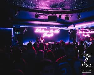 A crowded nightclub with neon pink signs on the walls. The sign in the background reads "Welcome to the Jungle." The room is dimly lit with an illuminated ceiling, and several people are gathered, facing towards the DJ booth where two DJs are performing. It's a scene you might explore in Uptown Waterloo. In the bottom right corner, there's a logo that reads "Zoo.
