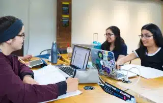 Three students, likely WUSA members, are studying together at a table. One has a laptop with colorful stickers, while another has notebooks and an open laptop displaying a document. The third student is writing in a notebook. They are all focused and working collaboratively with helpful supports around them.