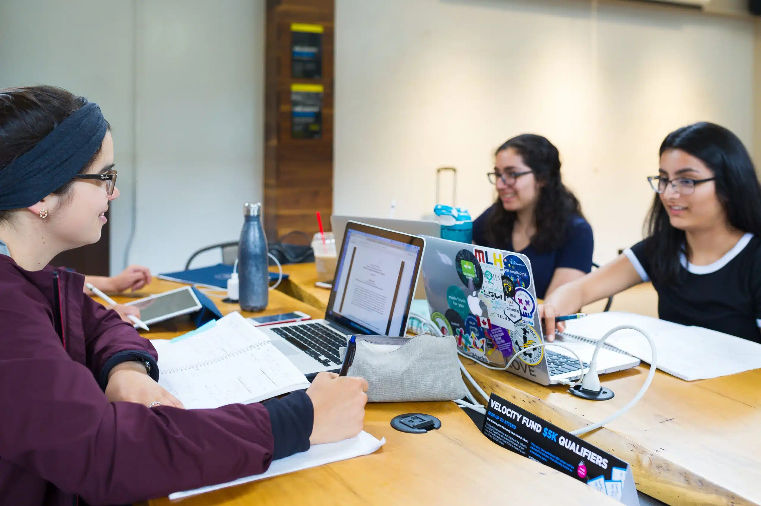 Three students, likely WUSA members, are studying together at a table. One has a laptop with colorful stickers, while another has notebooks and an open laptop displaying a document. The third student is writing in a notebook. They are all focused and working collaboratively with helpful supports around them.