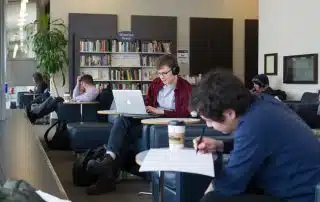 A group of people are in a cozy library lounge with bookshelves in the background. One person wears headphones and works on a laptop, while another writes on paper beside a coffee cup. Others are reading or working, creating a quiet and studious atmosphere that supports productivity.