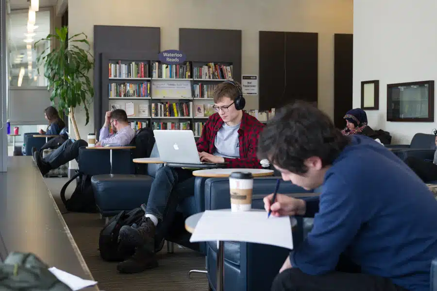 A group of people are in a cozy library lounge with bookshelves in the background. One person wears headphones and works on a laptop, while another writes on paper beside a coffee cup. Others are reading or working, creating a quiet and studious atmosphere that supports productivity.