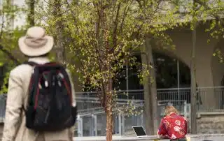 A person in a hat and backpack stands in the foreground, partially out of focus, while another person in a red floral shirt works on a laptop at an outdoor table under a tree, possibly analyzing new findings from a representative survey. The surrounding area has greenery and a building with large windows in the background.