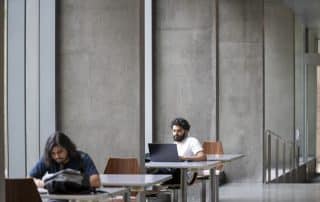 Two people are seated at separate tables in a modern, concrete-walled indoor space. One 2nd year student is working on a laptop, while the other is looking through papers, possibly for Reading Week tips. Both tables have wooden chairs, and large windows are present in the background.