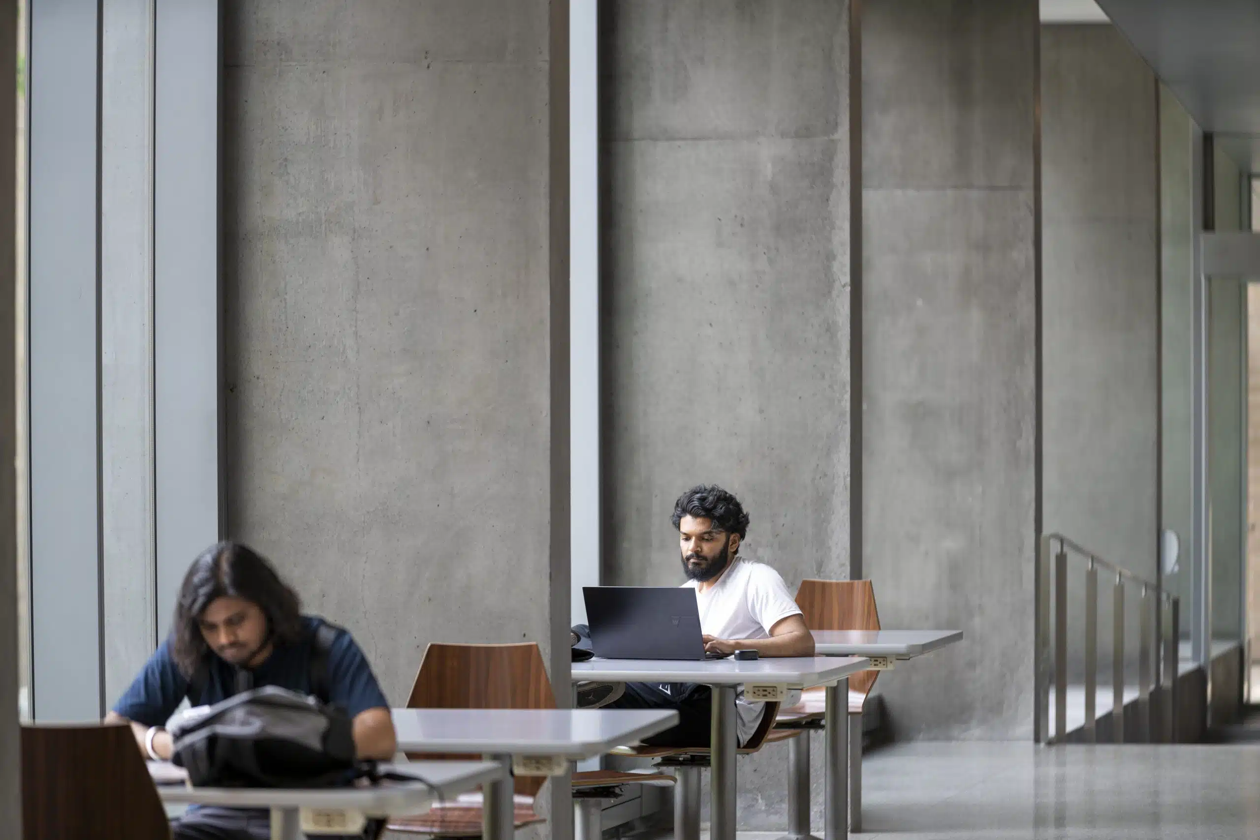 Two people are seated at separate tables in a modern, concrete-walled indoor space. One 2nd year student is working on a laptop, while the other is looking through papers, possibly for Reading Week tips. Both tables have wooden chairs, and large windows are present in the background.