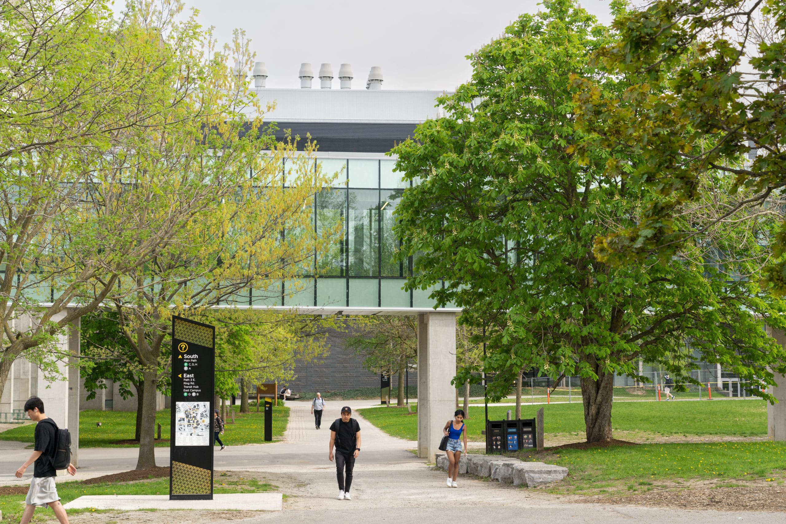 People walking along a pathway leading to a modern building with large glass windows. The path is surrounded by green grass and trees, some with budding leaves. A large information sign stands to the left featuring maps and directory details, perfect for 2nd year student tips during Reading Week.