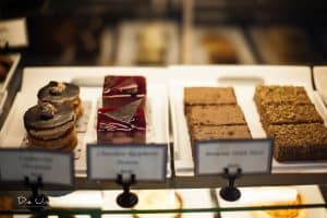 A display case in Uptown Waterloo showcases an assortment of desserts. From left to right, there are chocolate hazelnut pastries, raspberry mousse cakes with a chocolate garnish, brownies, and oat bars. Each dessert is neatly arranged on white trays with labels indicating their names, inviting you to explore every delightful option.