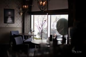 A cozy café in Uptown Waterloo with dim lighting featuring two small round tables with blue chairs near a large front window. A leafless branch decoration and a black-and-white photo of a ship adorn the space. The inviting ambiance is enhanced by a decorative chandelier, perfect for those looking to explore Waterloo's charming spots.