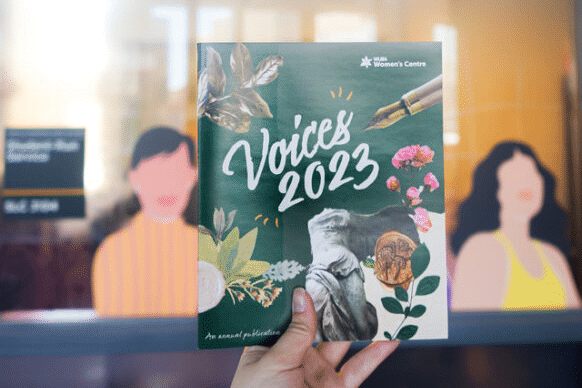 Image of a physical copy of Voices 2023 being held up in front of the Women's Centre Office