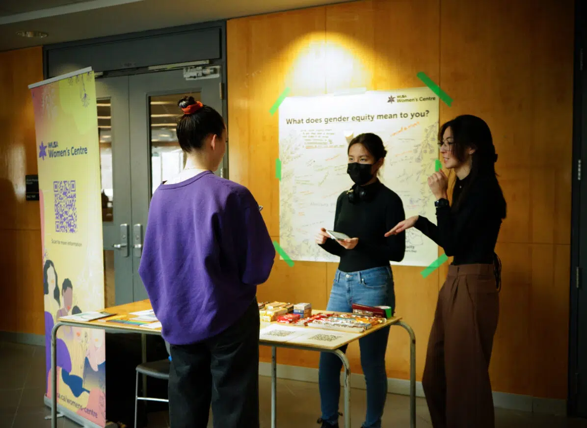 Image of two Women's Centre student volunteers giving a presentation and inviting a third student to participate in their booth activity.