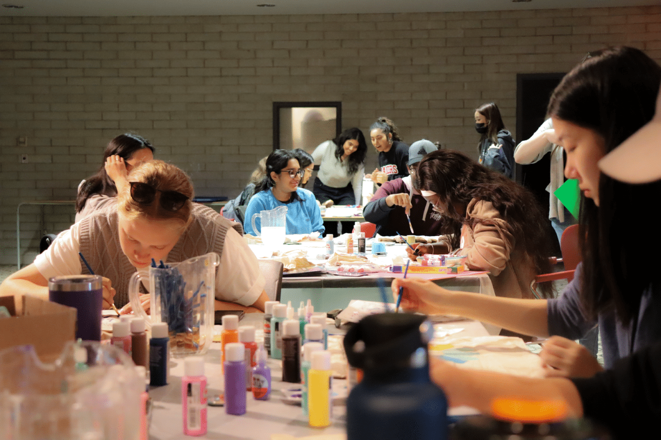 A group of people sits at tables engaged in arts and crafts activities. Various art supplies like paint, brushes, and paper are scattered on the tables. In the background, more people are interacting and working. The setting appears to be a well-lit indoor space.