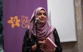 A woman wearing a pink headscarf and a black outfit is speaking into a microphone while holding a notebook. Behind her, a purple banner with a yellow flower logo and text that reads "The Women's" is partially visible. The setting, indicative of an indoor feminist community event, feels supportive.