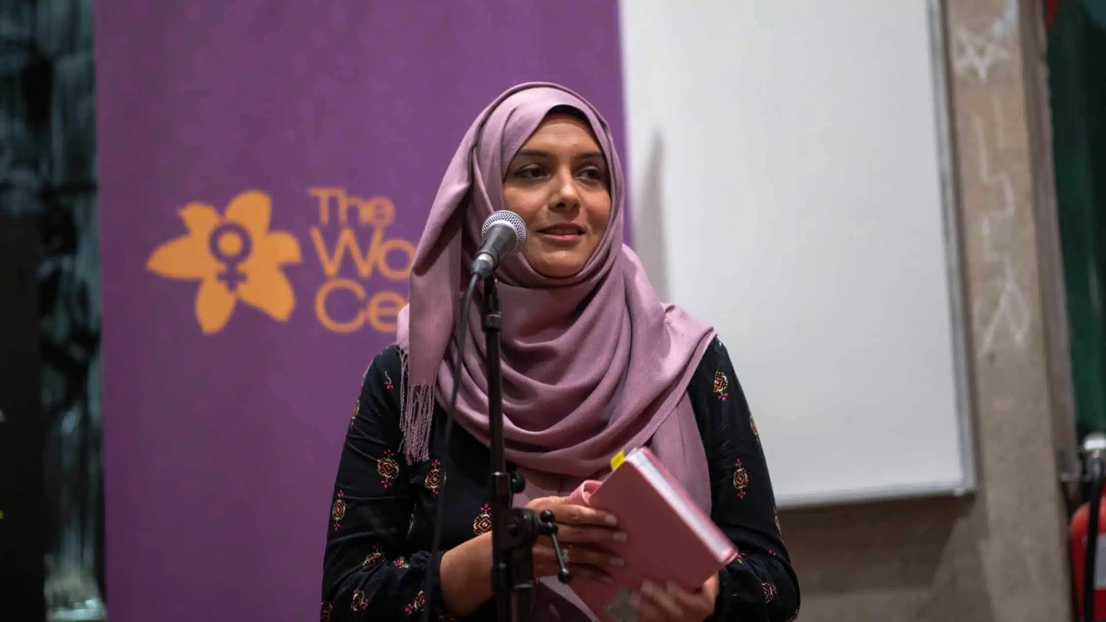 A woman wearing a pink headscarf and a black outfit is speaking into a microphone while holding a notebook. Behind her, a purple banner with a yellow flower logo and text that reads "The Women's" is partially visible. The setting, indicative of an indoor feminist community event, feels supportive.