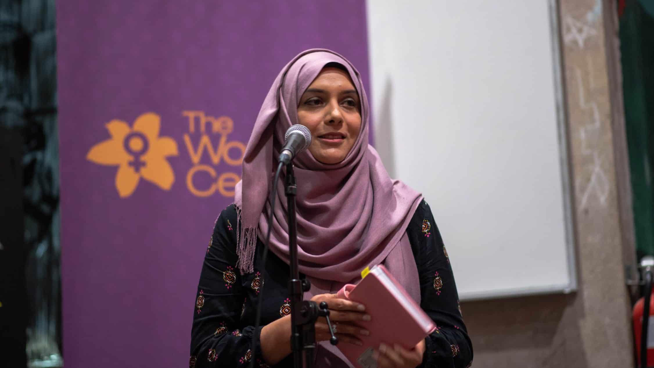 A woman wearing a purple hijab speaks into a microphone while holding a pink notebook. She stands in front of a purple banner with a yellow flower symbol and the text "The Women's Center," embodying the spirit of the feminist support community gathered there.