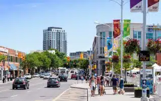 A bustling urban street scene on a sunny day, with pedestrians exploring the sidewalk and vehicles driving on the road. Colorful banners hang from streetlights, and a tall modern building is visible in the background. Shops and greenery line both sides of the street in Uptown Waterloo.