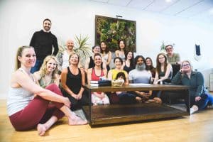 A group of fifteen people of diverse age and gender smile while posing for a photo in a yoga studio. They are gathered around a wooden table, with a green plant wall in the background. A baby sits on the table, surrounded by the cheerful group, capturing the spirit of Uptown Waterloo exploration.