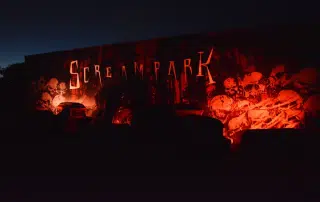 A dark amusement park set with eerie red lighting, featuring a large sign that reads "Scream Park" and displaying numerous skulls on the wall. Various objects and decorations enhance the spooky atmosphere, making it a must-visit for Halloween activities in the KW Region.