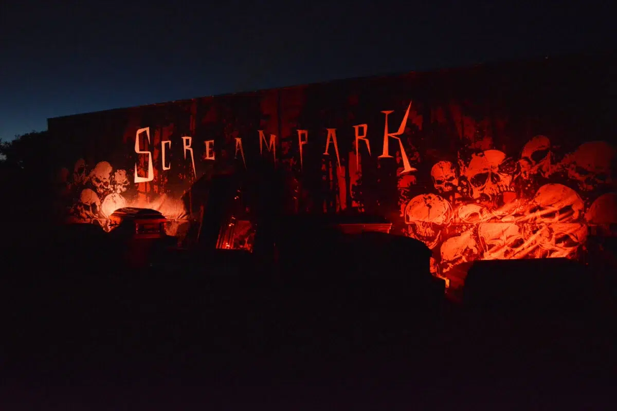 A dark amusement park set with eerie red lighting, featuring a large sign that reads "Scream Park" and displaying numerous skulls on the wall. Various objects and decorations enhance the spooky atmosphere, making it a must-visit for Halloween activities in the KW Region.