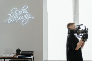 A person in black clothing and gloves operates a large professional camera inside a well-lit room with a white neon sign that reads "Sasha tattooing" on the wall. A table with various items is visible in the corner of the room, capturing one of Uptown Waterloo's unique attractions.