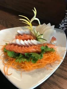 A beautifully presented plate of sashimi features neatly arranged slices of salmon and white fish on a bed of shredded carrots and green lettuce. The dish, perfect for those looking to explore the culinary delights of Uptown Waterloo, is garnished with thinly sliced lemon zest and cucumber, creating an elegant and appetizing display.