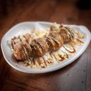 A white plate with a curved design holds four takoyaki (Japanese octopus balls) drizzled with mayonnaise and okonomiyaki sauce, garnished with bonito flakes and chopped parsley, placed on a wooden table—an enticing dish to explore Uptown Waterloo.