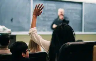 A student with long dark hair raises their hand to participate in the classroom. The educator, standing in front of a chalkboard with writing on it, points towards the student. Surrounding students are seated, facing the front, all eager to contribute to their educational journey.