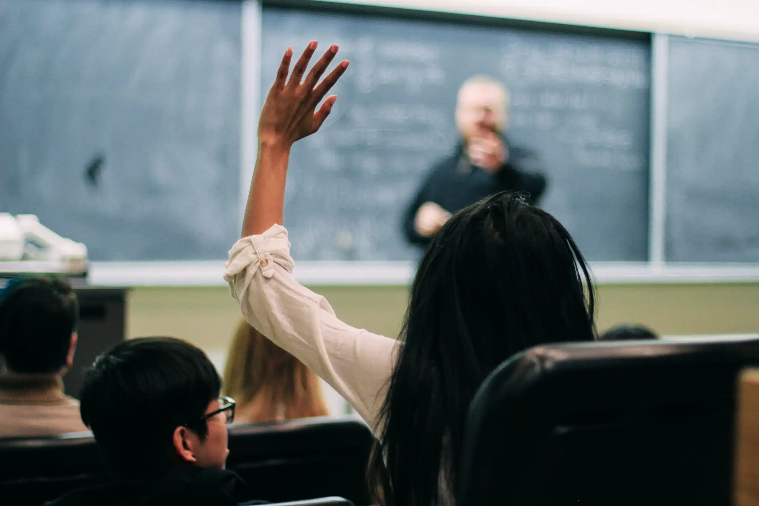 A student with long dark hair raises their hand to participate in the classroom. The educator, standing in front of a chalkboard with writing on it, points towards the student. Surrounding students are seated, facing the front, all eager to contribute to their educational journey.