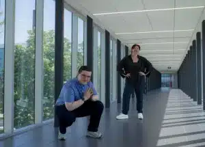 Two people are posing in an indoor corridor with large windows. One person is crouched with hands together, while the other stands behind with hands on hips. The corridor with its tiled floor offers a view of trees through the windows, reminiscent of the strategic planning often seen during political campaigns.