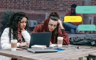 Two individuals sit at a wooden table outdoors, focused on a laptop during an OUSA consultation. Both appear engaged in their work, surrounded by books and coffee cups. Three chat bubbles hover near the image, indicating a conversation. A brick wall and another person are visible in the background.