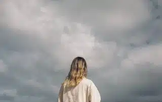 A person with shoulder-length hair, wearing a beige jacket, is standing outdoors and looking up at a cloudy sky, perhaps reflecting on seasonal depression. The back of the person is visible, and the overcast sky occupies the majority of the background.