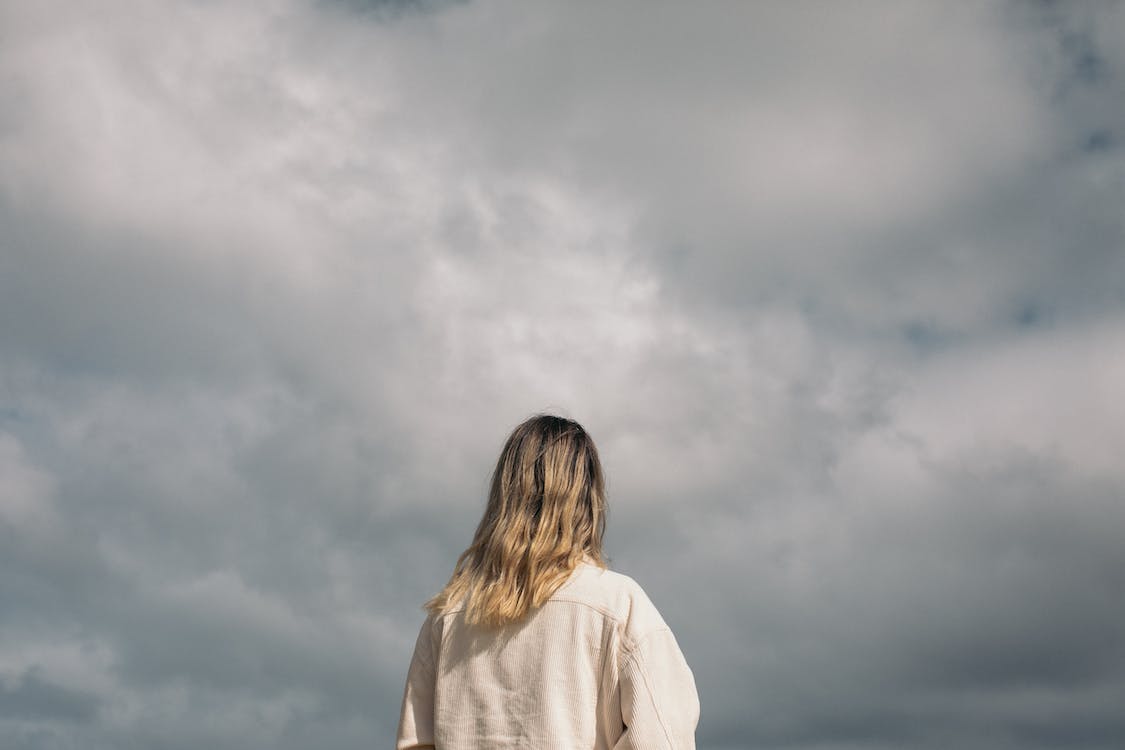 A person with shoulder-length hair, wearing a beige jacket, is standing outdoors and looking up at a cloudy sky, perhaps reflecting on seasonal depression. The back of the person is visible, and the overcast sky occupies the majority of the background. A person with shoulder-length hair, wearing a beige jacket, is standing outdoors and looking up at a cloudy sky, perhaps reflecting on seasonal depression. The back of the person is visible, and the overcast sky occupies the majority of the background.