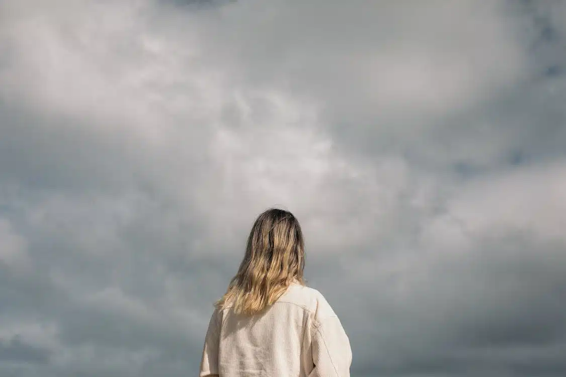 A person with shoulder-length hair, wearing a beige jacket, is standing outdoors and looking up at a cloudy sky, perhaps reflecting on seasonal depression. The back of the person is visible, and the overcast sky occupies the majority of the background.