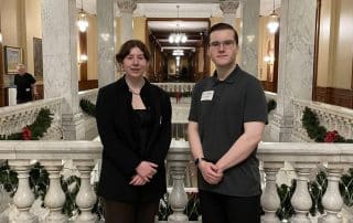 Two people, including the WUSA President, stand side by side in a grand marble hallway with intricate railings. They are dressed in formal attire, with marble pillars and Christmas decorations visible in the background. Both are facing the camera and smiling gently.