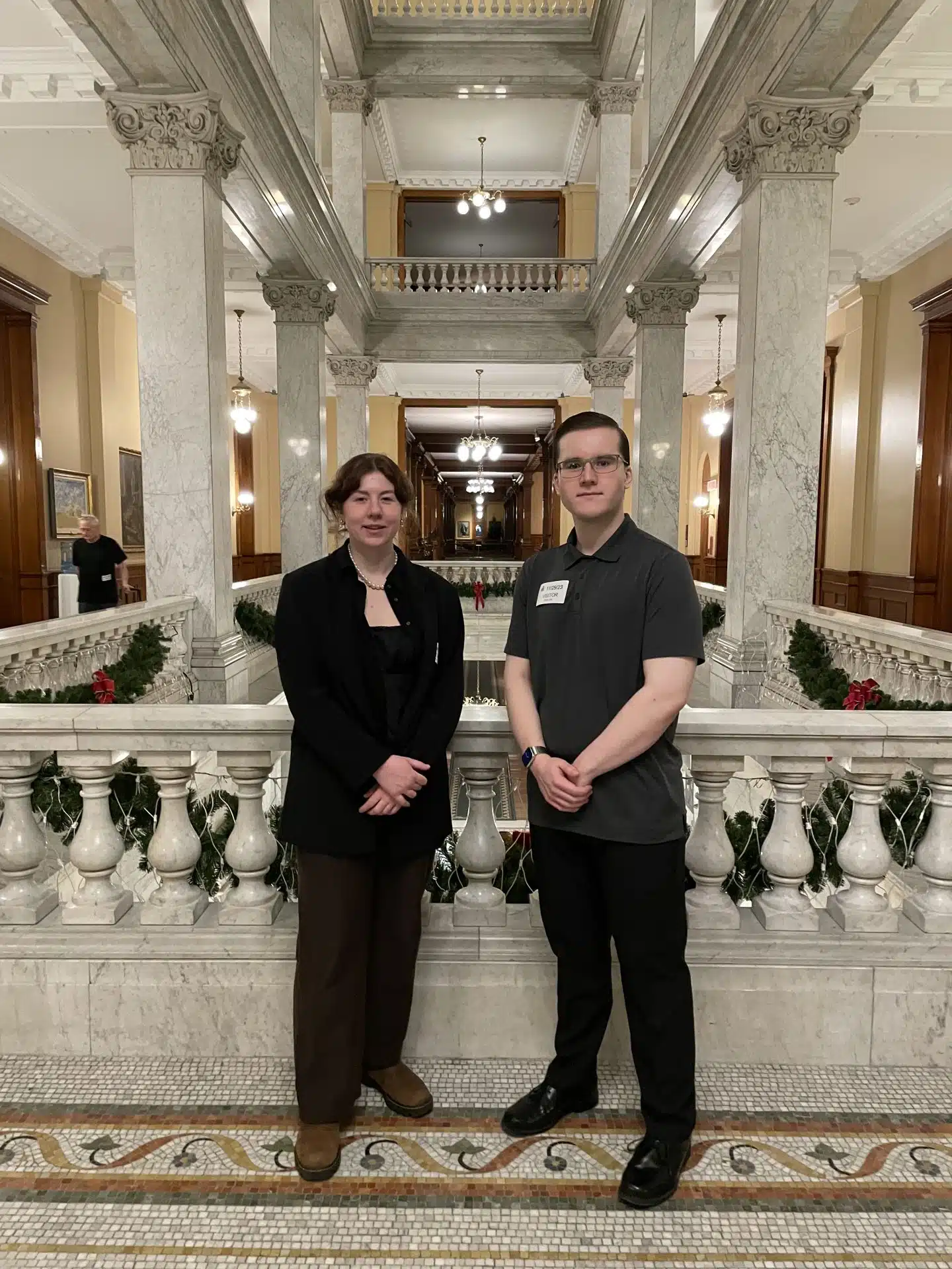 Two people, including the WUSA President, stand side by side in a grand marble hallway with intricate railings. They are dressed in formal attire, with marble pillars and Christmas decorations visible in the background. Both are facing the camera and smiling gently.