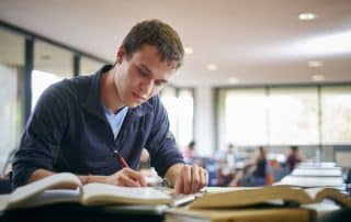 A young UW student with short brown hair, wearing a navy blue jacket over a light blue shirt, sits at a desk in the library, writing Final Exam Tips in a notebook. Open books are spread out in front of him, and large windows are in the background.