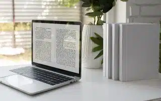 A laptop displaying text from an open e-book is placed on a white desk, promoting ways to save money on books. Next to the laptop are three blank white books stacked upright. A potted plant is in the background near a window with blinds, casting gentle natural light into the room.