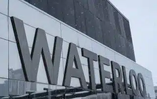 Outdoor signage displaying the large metallic word "WATERLOO" on the facade of a modern building, with winter snow falling lightly in the background. The scene captures the beauty of Waterloo, perfect for a UW Photography Club session.