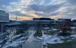 A scenic view of a modern campus at sunset in Waterloo, with buildings, pathways, and patches of winter snow on the ground. The sky is filled with dramatic, wispy clouds illuminated by the setting sun, casting a serene atmosphere over the scene. Trees are scattered around the area, perfect for photography.