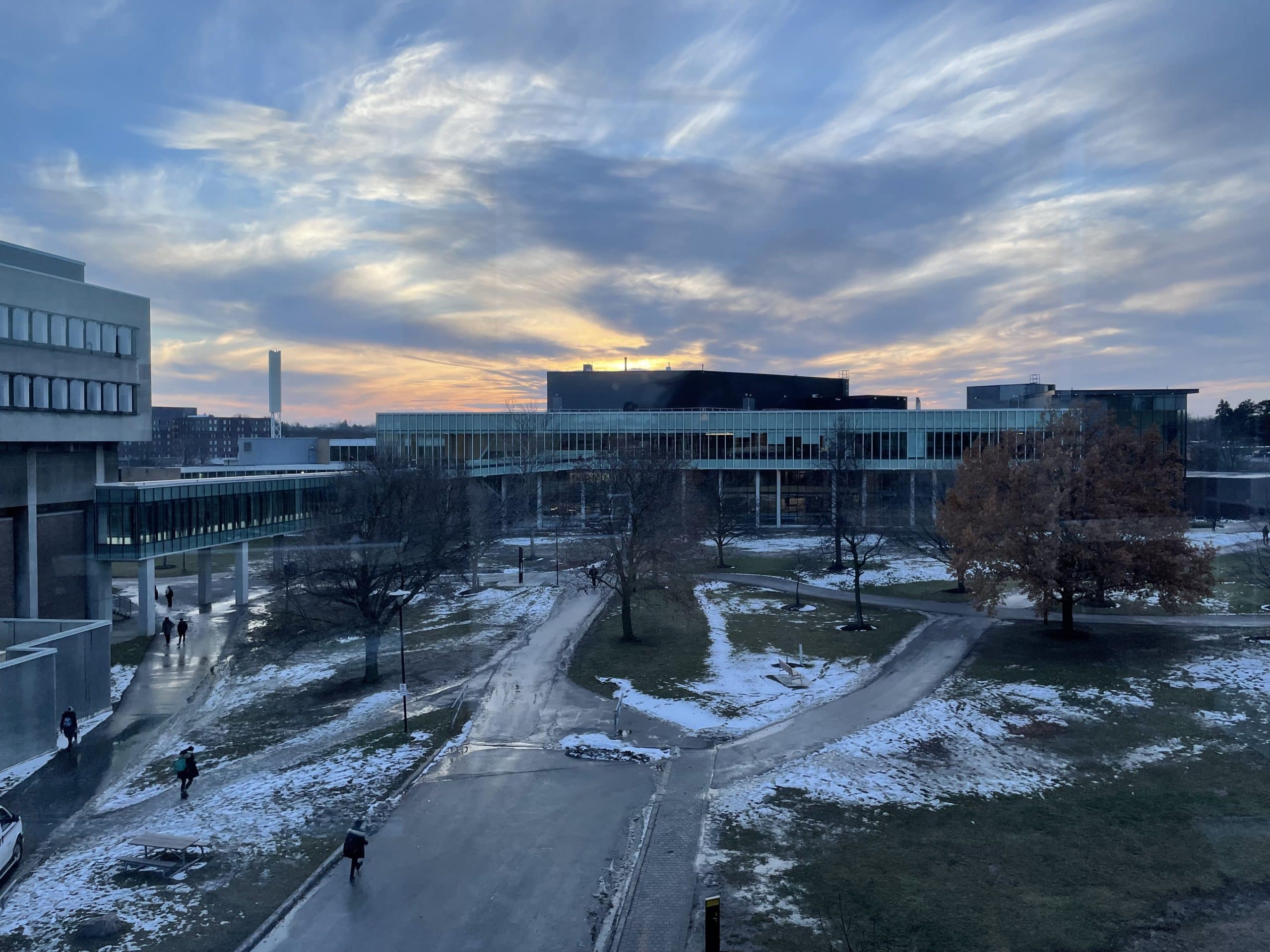 A scenic view of a modern campus at sunset in Waterloo, with buildings, pathways, and patches of winter snow on the ground. The sky is filled with dramatic, wispy clouds illuminated by the setting sun, casting a serene atmosphere over the scene. Trees are scattered around the area, perfect for photography.