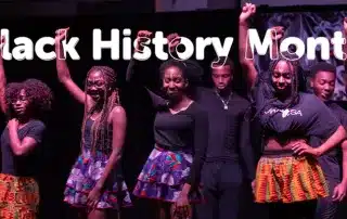 A group of people wearing colorful skirts and black tops stand on a stage with their fists raised in celebration. The text "Black History Month: Writing Our History" is prominently displayed above them. The dark background emphasizes the vibrant attire and the powerful message of their raised fists.