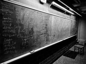 The room, dimly lit with fluorescent lights, showcases a large black chalkboard covered in mathematical and electrical engineering equations, notations, and diagrams. Two wooden chairs are positioned near the board. This scene could easily be a contender in February Photo Contest's "Favourite Study Spot" category.