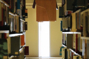 A library aisle with shelves filled with books on both sides. At the end of the aisle, light shines through a tall window, illuminating the path—a setting that won praise in our February Photo Contest. A wooden rectangular structure hangs from the ceiling in the center near the window.