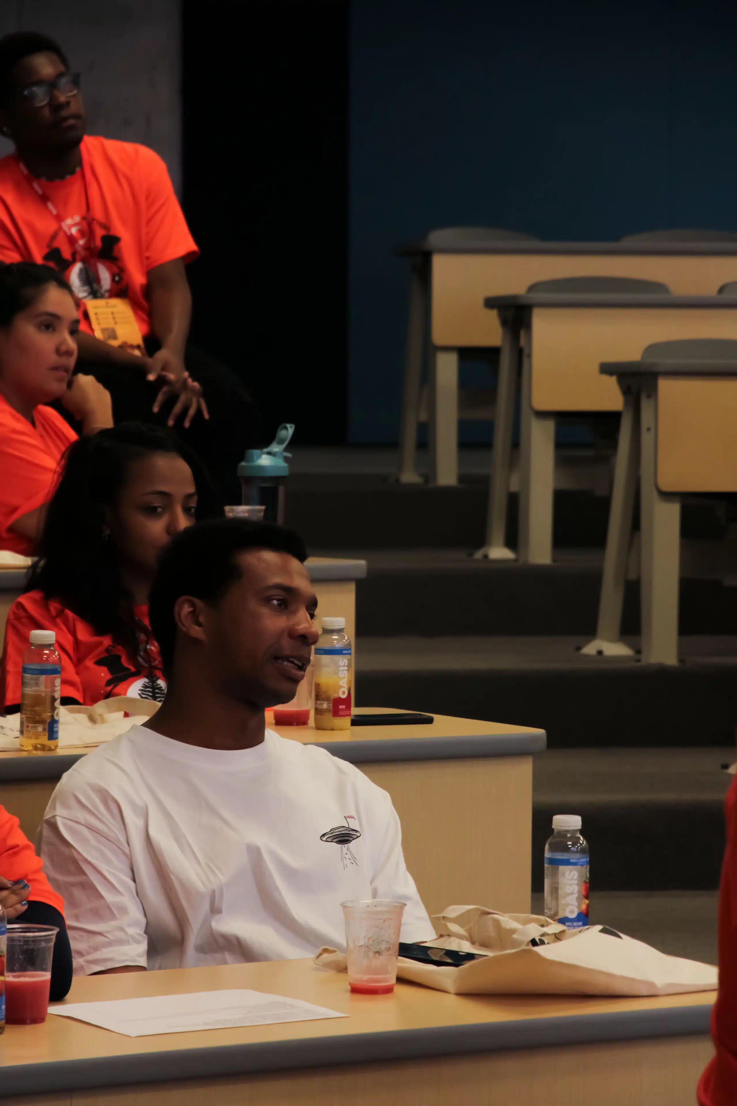 A group of people in a classroom setting, attentively listening to someone. The focus is on a person wearing a white shirt seated at a desk, surrounded by others in red shirts. Desks are adorned with water bottles and papers. The environment has a student-run vibe, fostering learning and collaboration.
