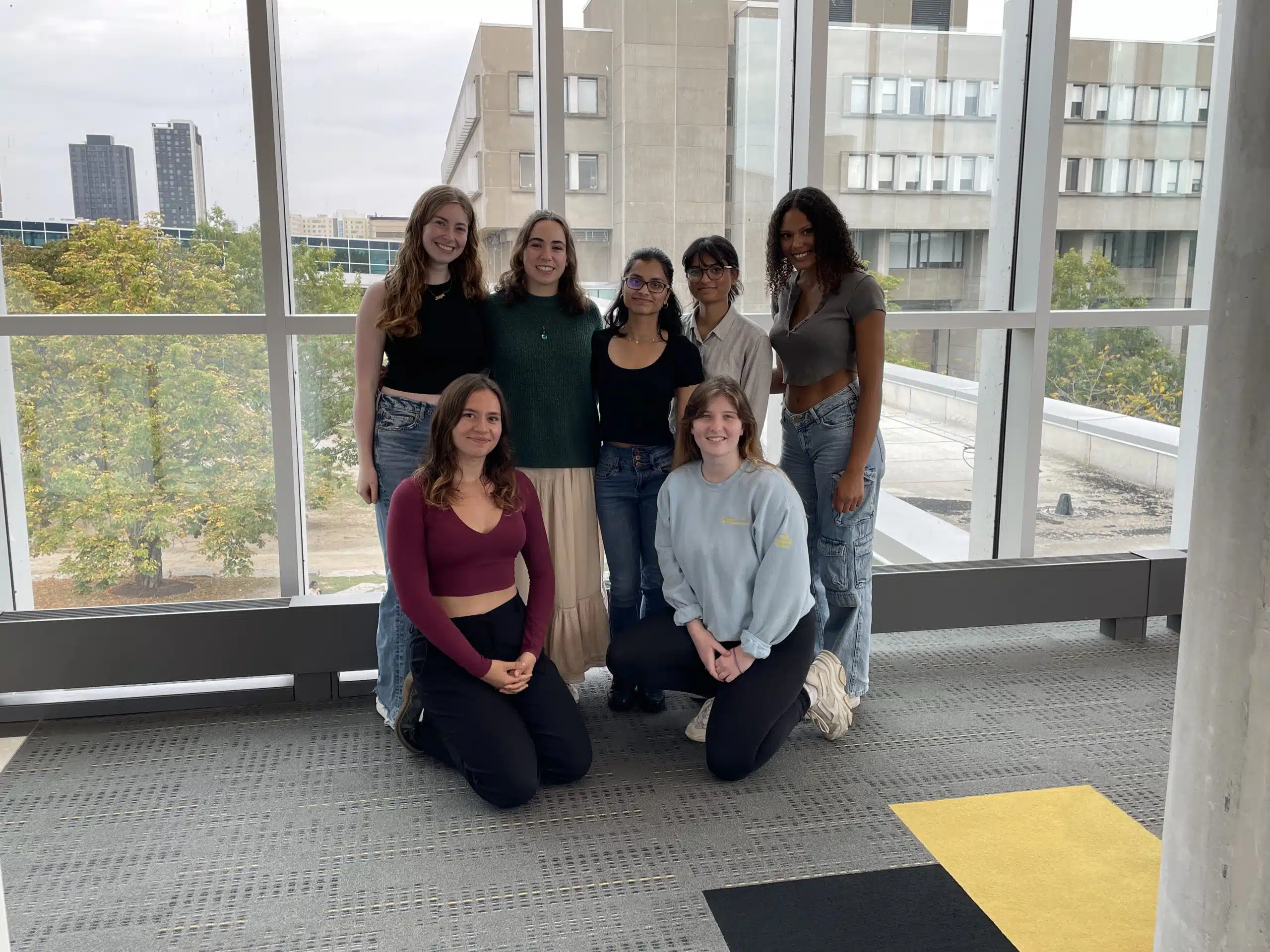 A group of seven diverse individuals, involved in Student-Run Services, pose for a photo in front of large windows with a cityscape in the background. Five stand while two kneel on yellow and black mats, set indoors in a modern building with gray flooring.