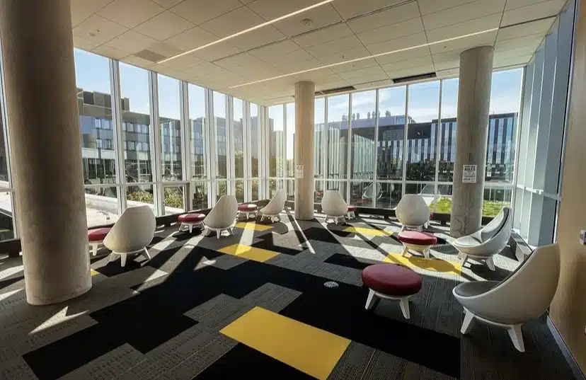 A modern indoor seating area with large floor-to-ceiling windows, natural light streaming in. White, curved chairs with red and white cushions are arranged around a patterned carpet with black and yellow squares. This could easily be your favourite study spot or an entry for the February Photo Contest, with tall buildings visible outside.