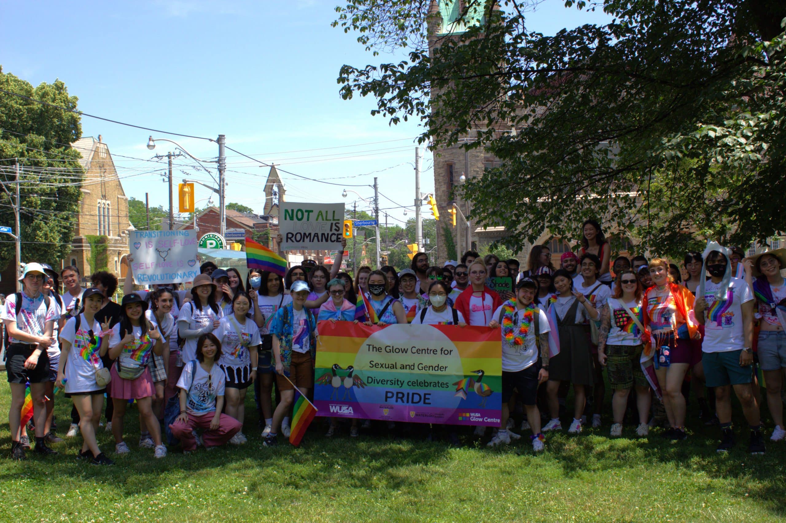 A large group of people poses together outdoors while holding a rainbow banner that reads, “The Glow Centre for Sexual and Gender Diversity celebrates PRIDE.” As a student-run initiative, some participants hold rainbow flags and signs. Trees and buildings are visible in the background, highlighting the community services. A large group of people poses together outdoors while holding a rainbow banner that reads, "The Glow Centre for Sexual and Gender Diversity celebrates PRIDE." As a student-run initiative, some participants hold rainbow flags and signs. Trees and buildings are visible in the background, highlighting the community services.