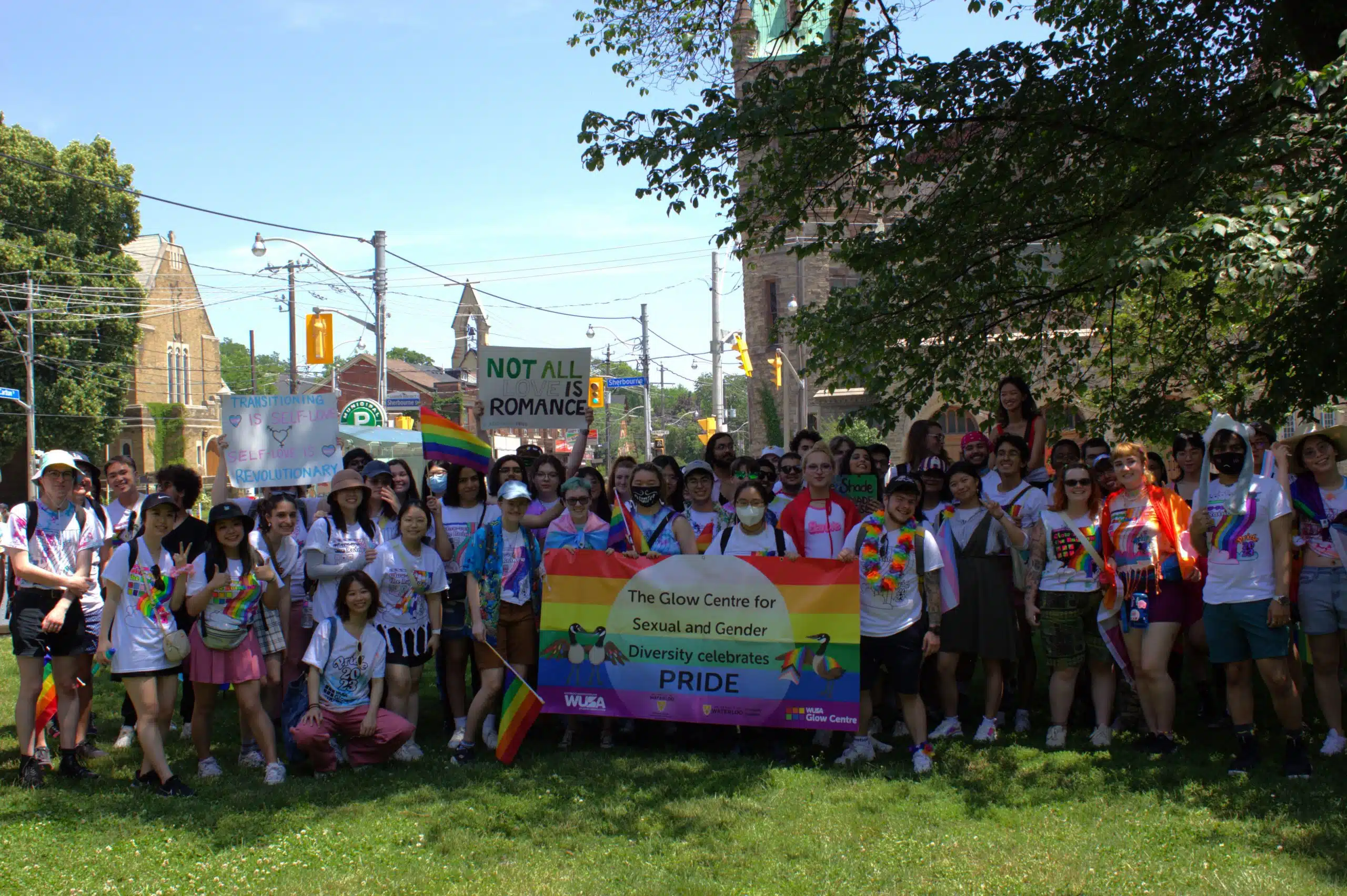 A large group of people poses together outdoors while holding a rainbow banner that reads, "The Glow Centre for Sexual and Gender Diversity celebrates PRIDE." As a student-run initiative, some participants hold rainbow flags and signs. Trees and buildings are visible in the background, highlighting the community services.
