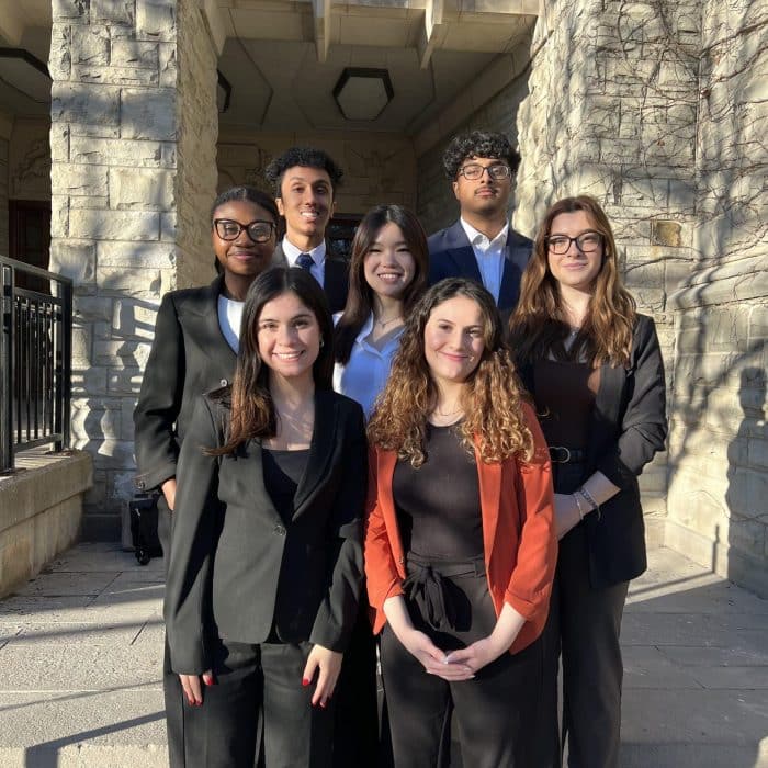 A group of seven people, four women and three men, all dressed in business attire, stand outside a stone building. In the front row are two women, the middle row features three individuals, and the back row consists of two men. They are posing and smiling for the camera following a successful event organized by the Mock Trial Club - Donation.